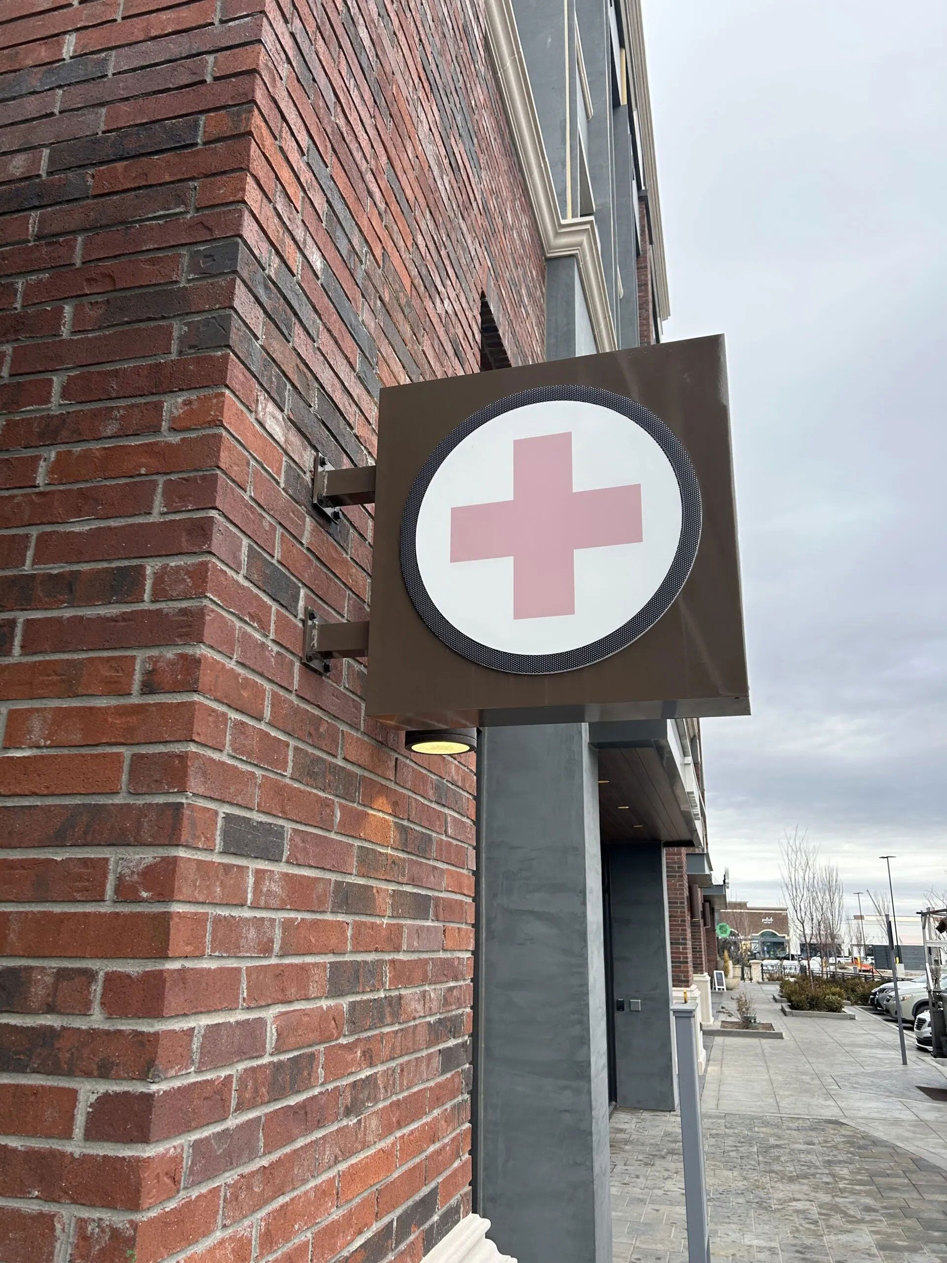 A brown, square sign with a white circle and red cross mounted to a red brick building on a city street.