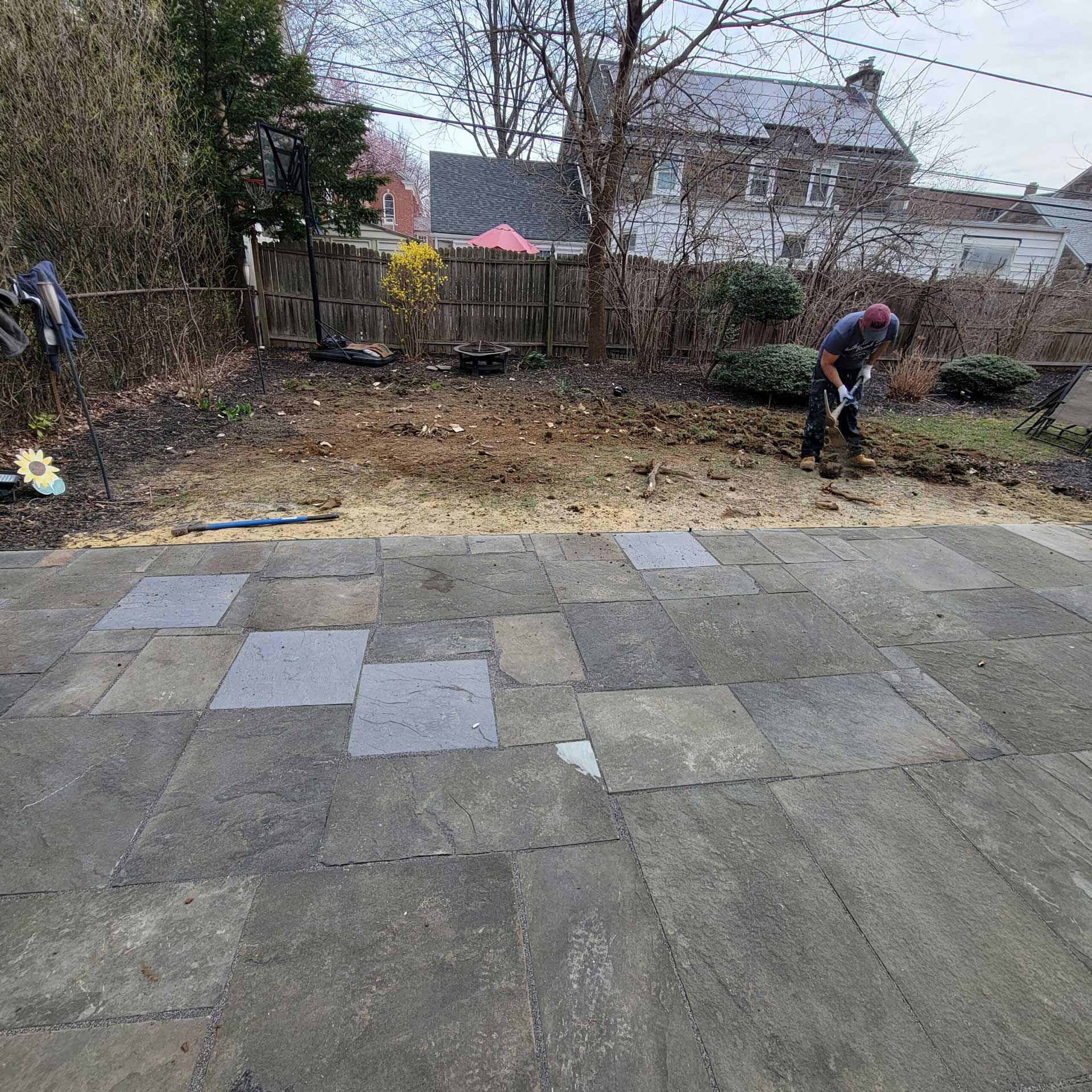 Outdoor patio with scattered paving stones, two people working in a backyard, and houses in the background