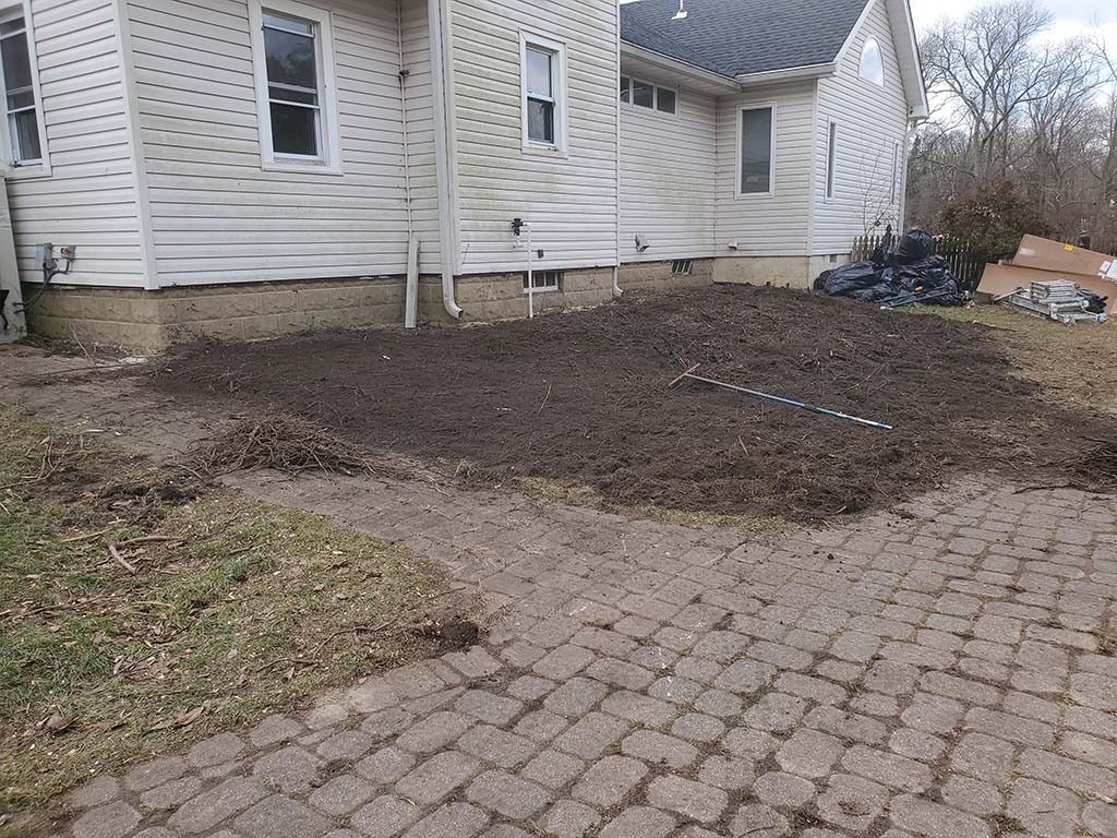 Freshly landscaped backyard beside a beige house, with bare soil and a brick patio in the foreground