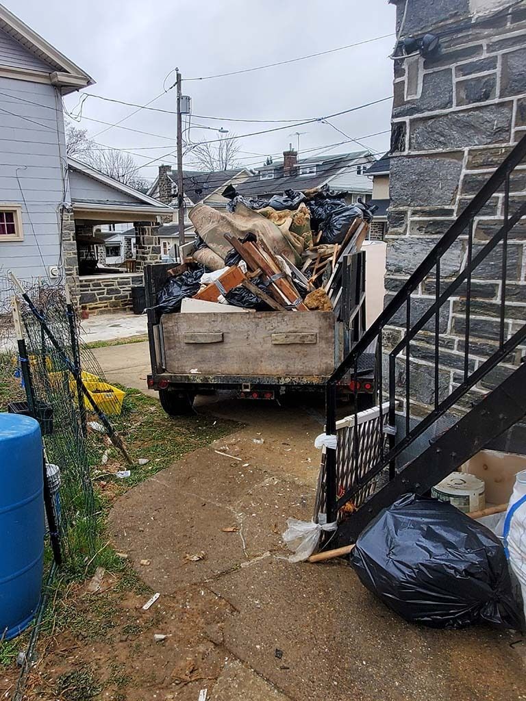 Dumpster full of debris in a narrow alley beside a stone house and metal stairs.