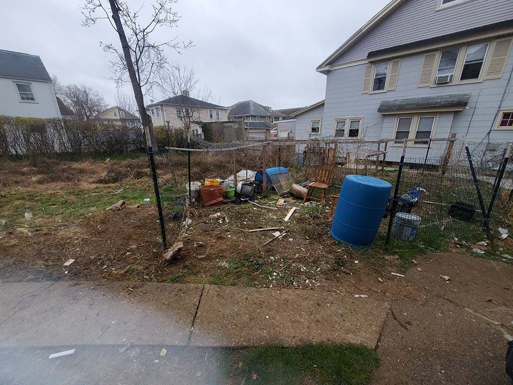 Rainy yard beside a gray house, with scattered debris, a blue barrel, and a bare tree.