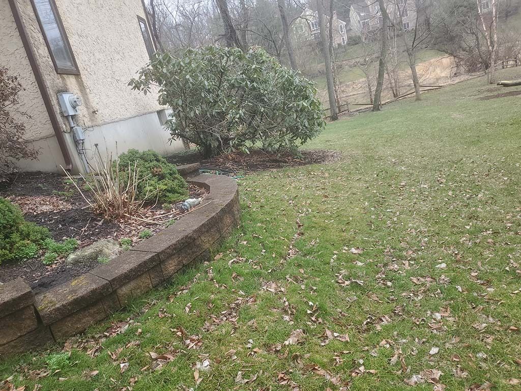 Side yard with a curved brick planter, shrubs, and a grassy lawn beside a house and wooded background