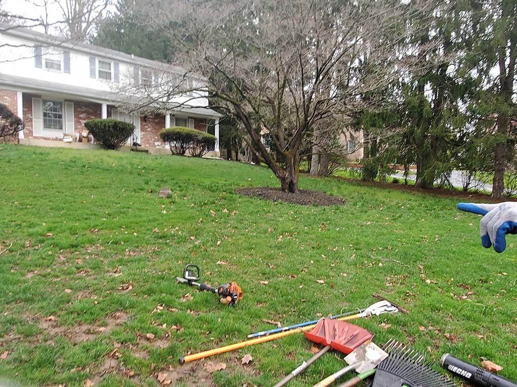 Yard with scattered tools on the grass in front of a townhouse, including a rake, shovel, and leaf blower.
