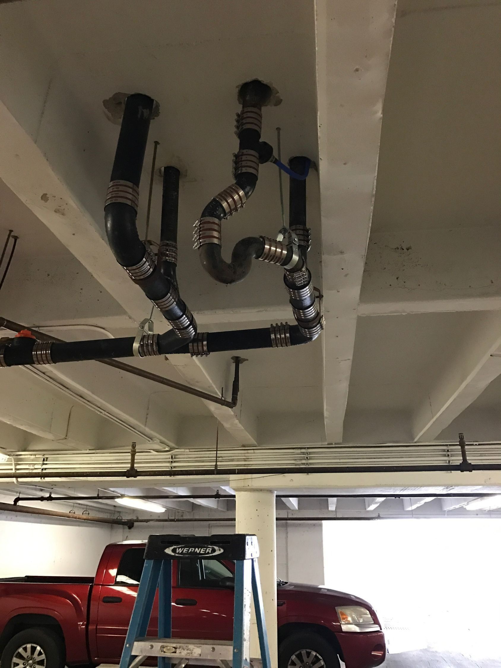 Black plumbing pipes attached to a white ceiling in a parking garage; a red truck and a ladder are visible below.