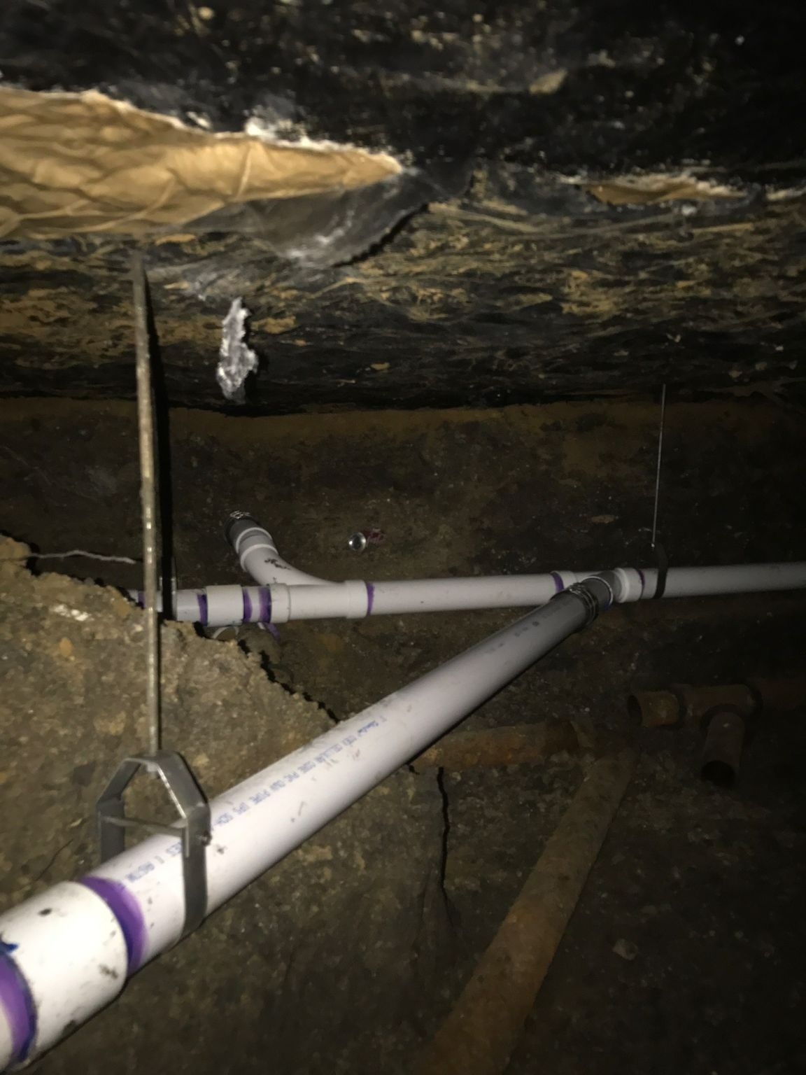 Pipes secured in a dark crawlspace; black ceiling, white pipes, and wooden supports.