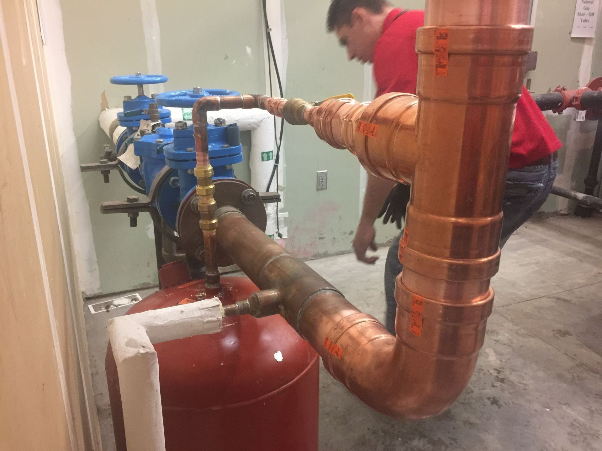 Plumber working on copper pipes, near a blue valve and red tank.