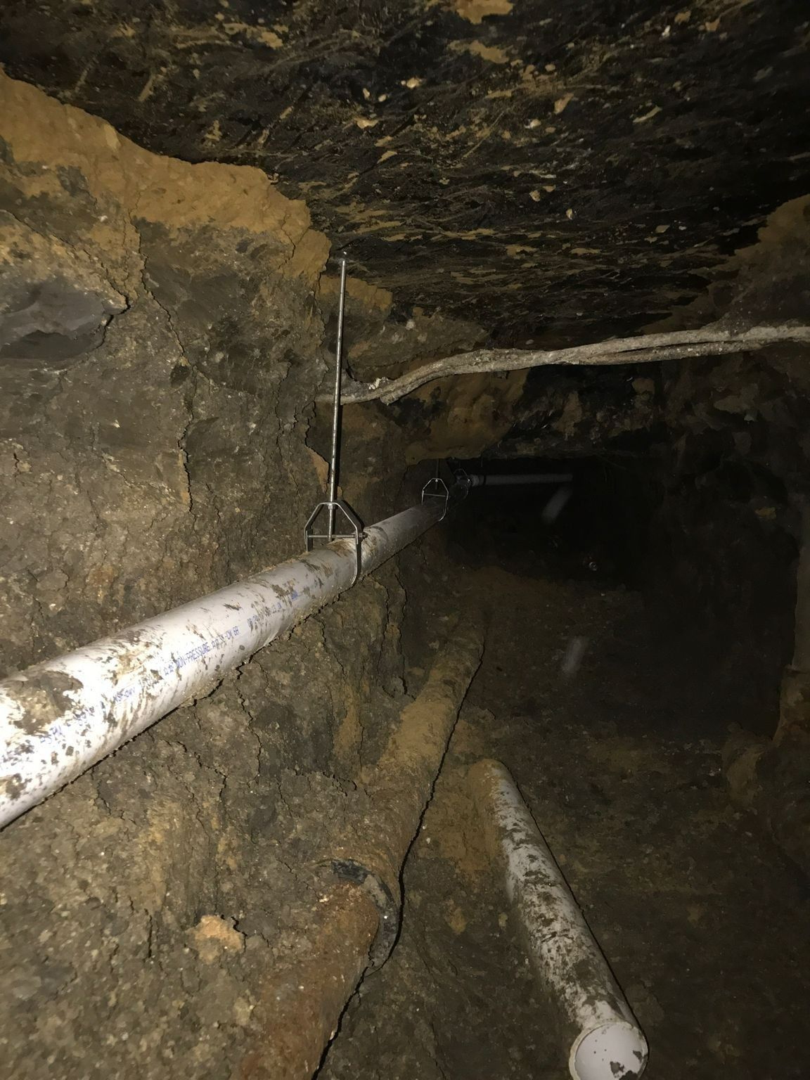 Tunnel with white pipes, suspended from the dark, dirt ceiling and walls.