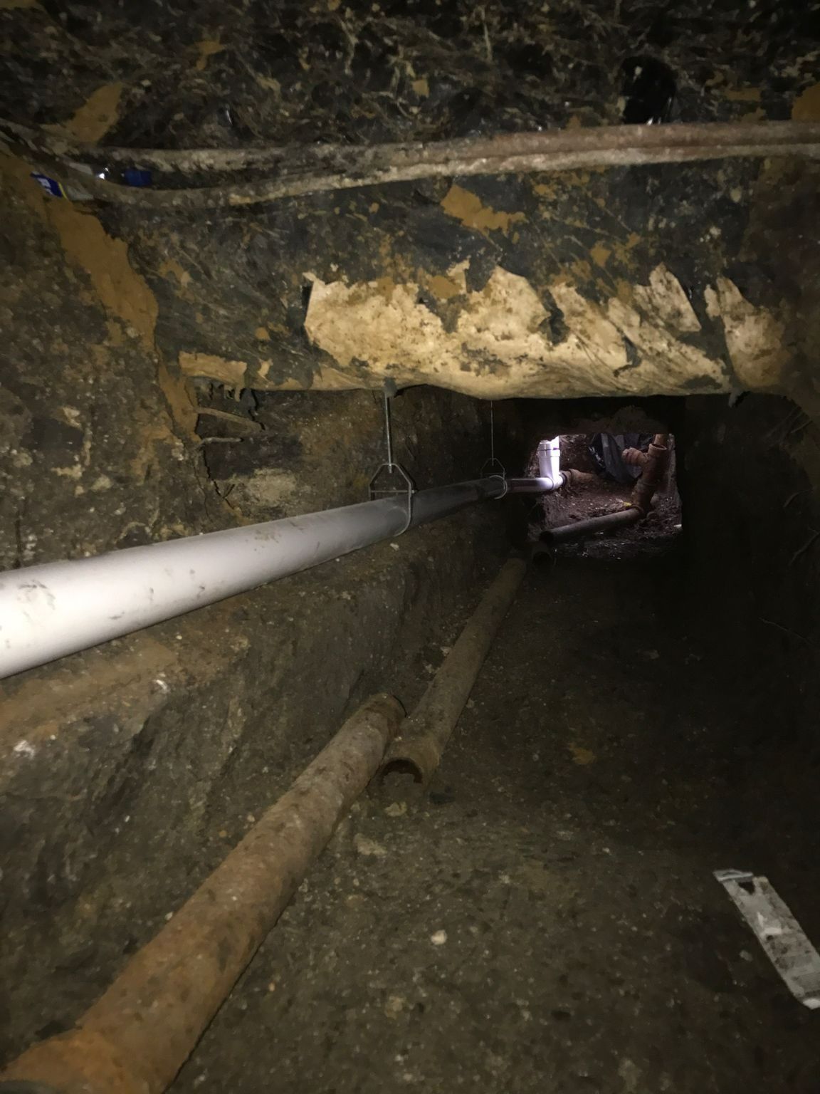A narrow, dark crawl space with PVC and rusty pipes, likely under a building foundation.