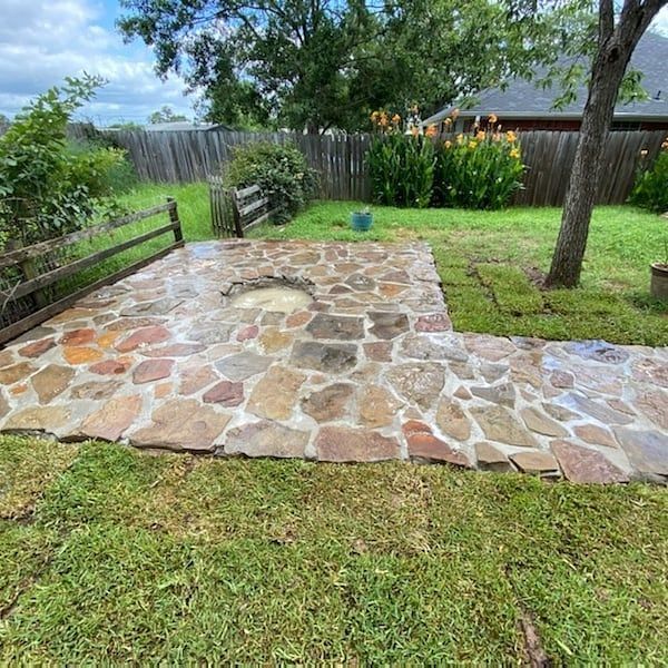 A stone patio with a fire pit in the middle of a lush green yard.
