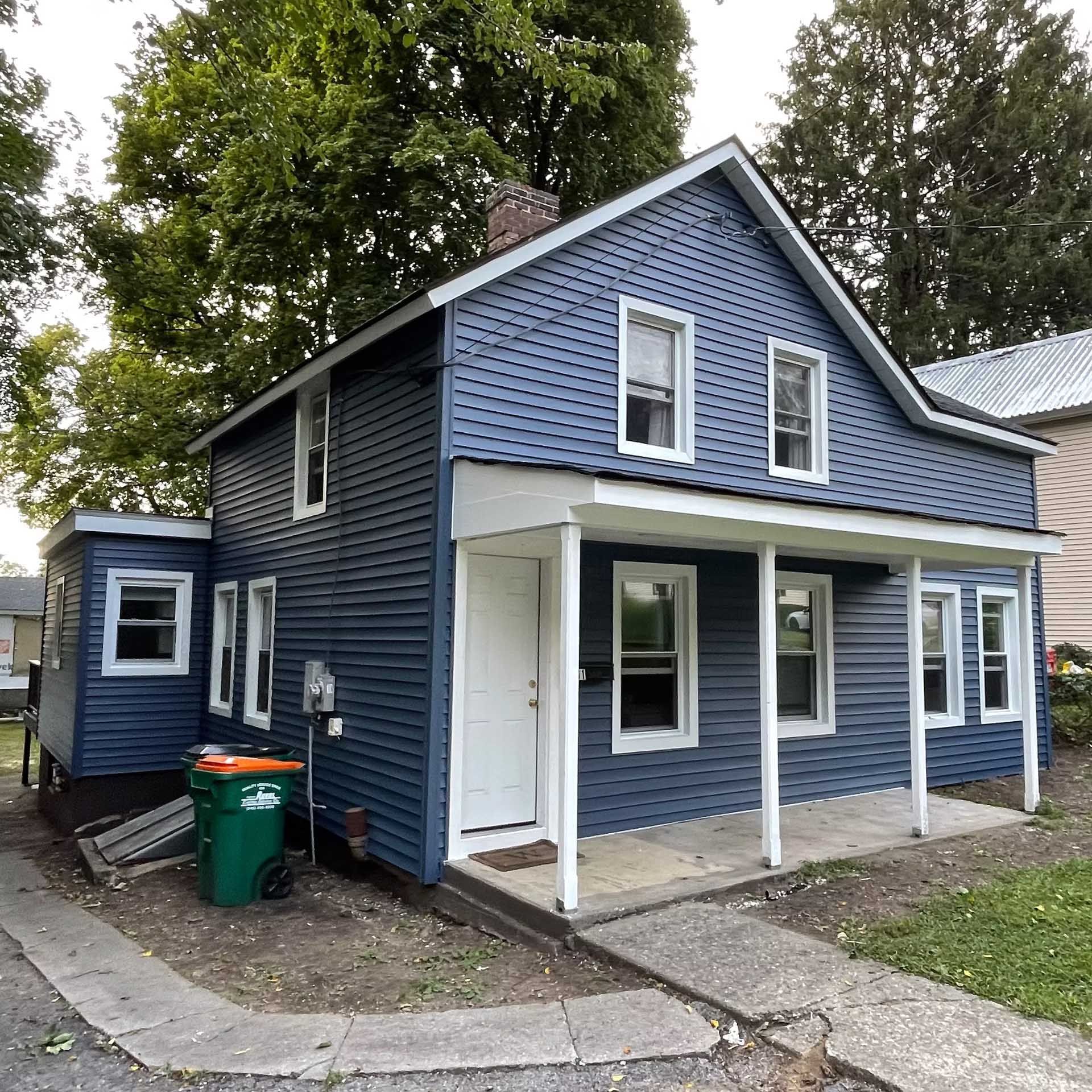 Two-story blue house with white trim and a porch, with trash can in front.