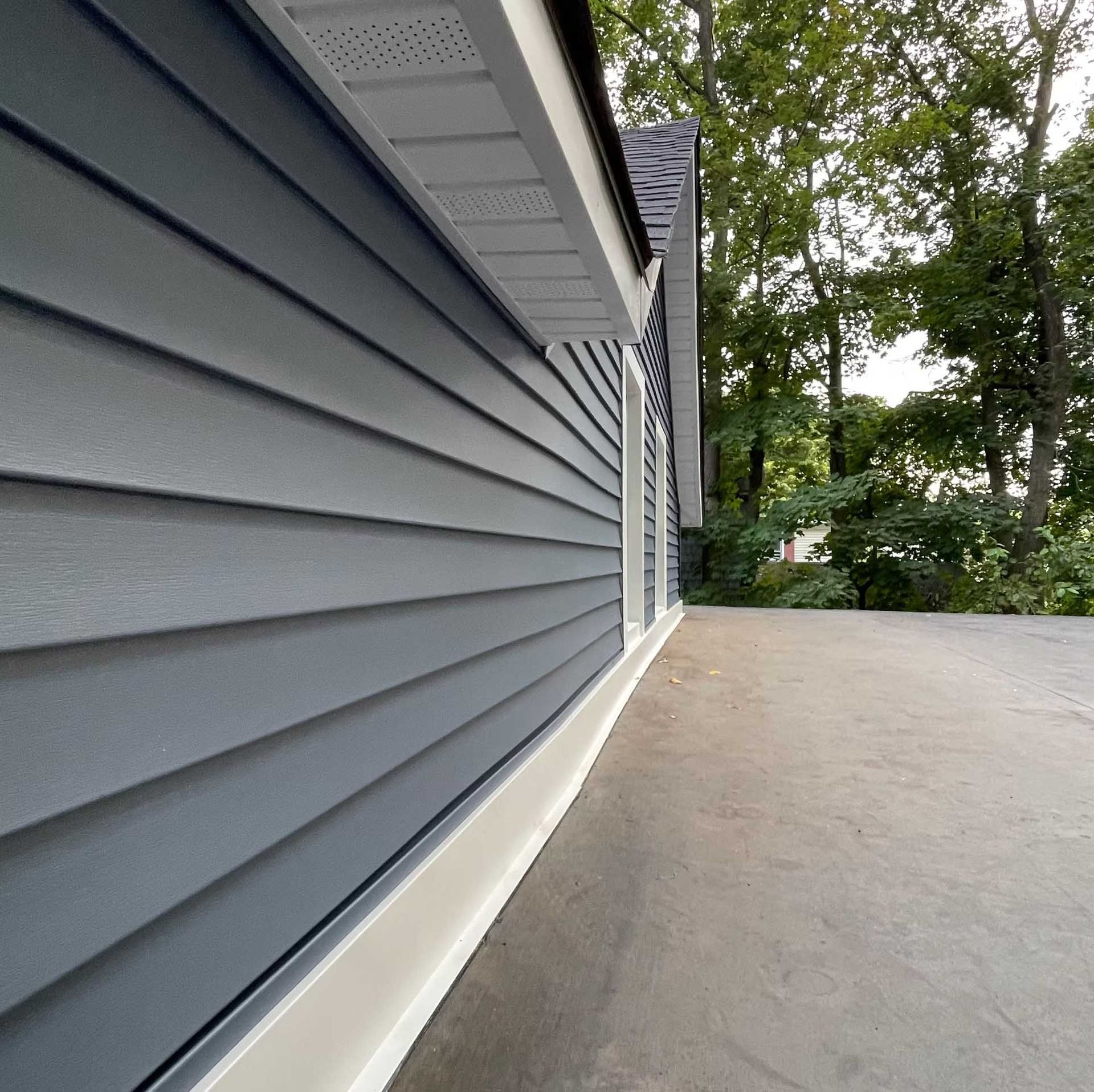 Gray house siding with white trim, angled view, concrete surface, trees in background.