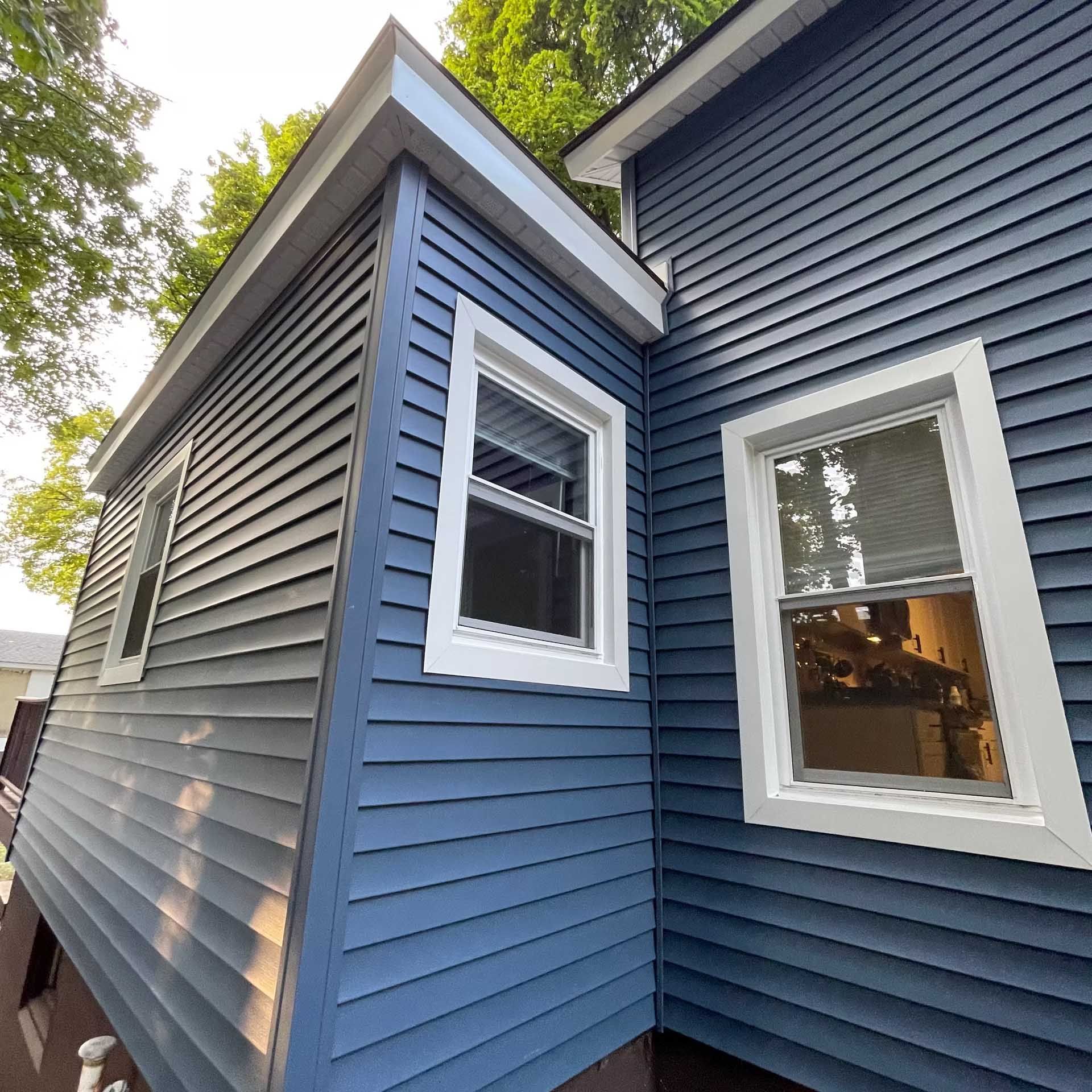 Exterior of a house with blue and gray siding, white window frames, and a light blue roof against greenery.