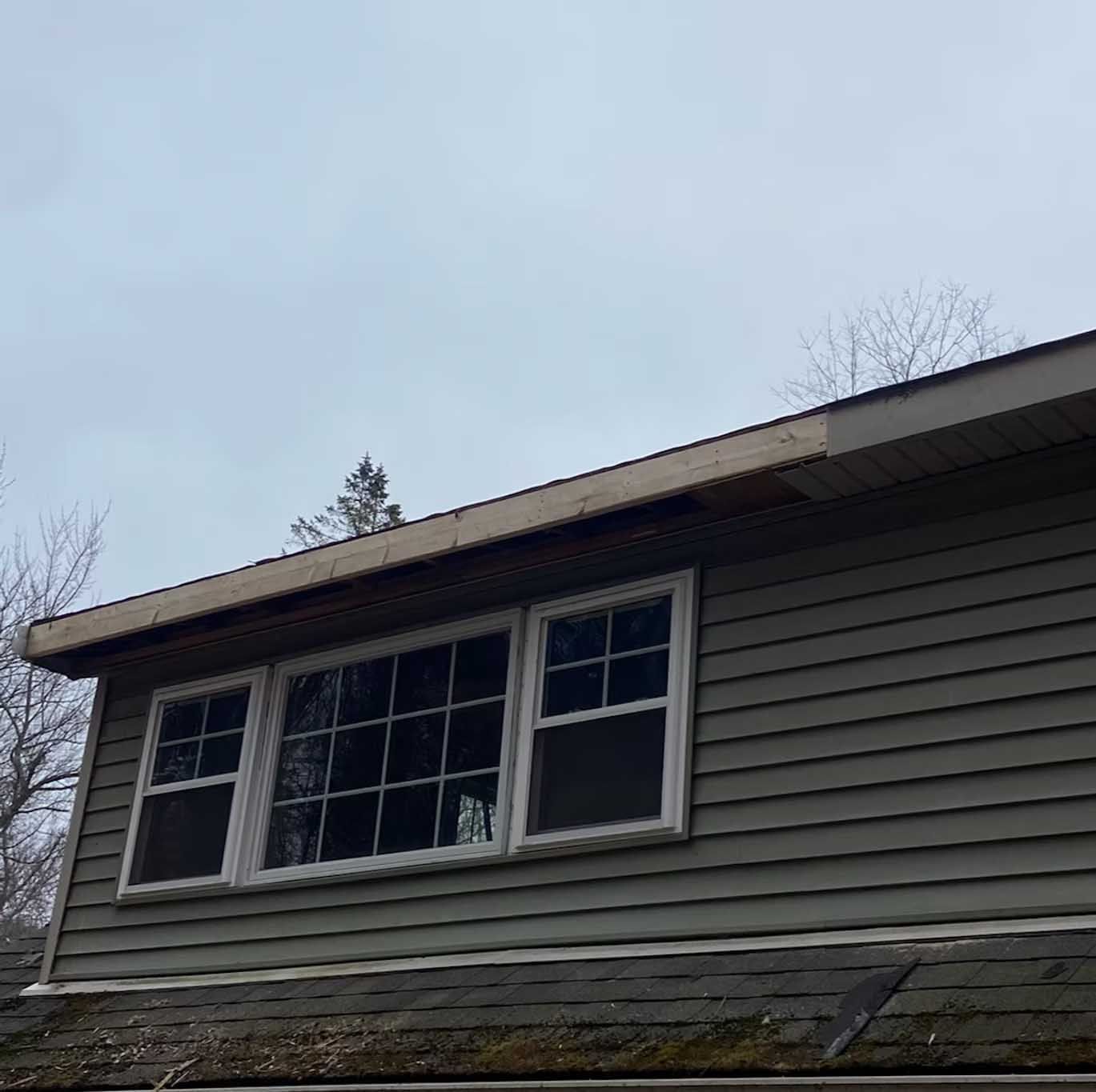 Exterior view of a house with gray siding, windows, and an overhanging roof under a cloudy sky.