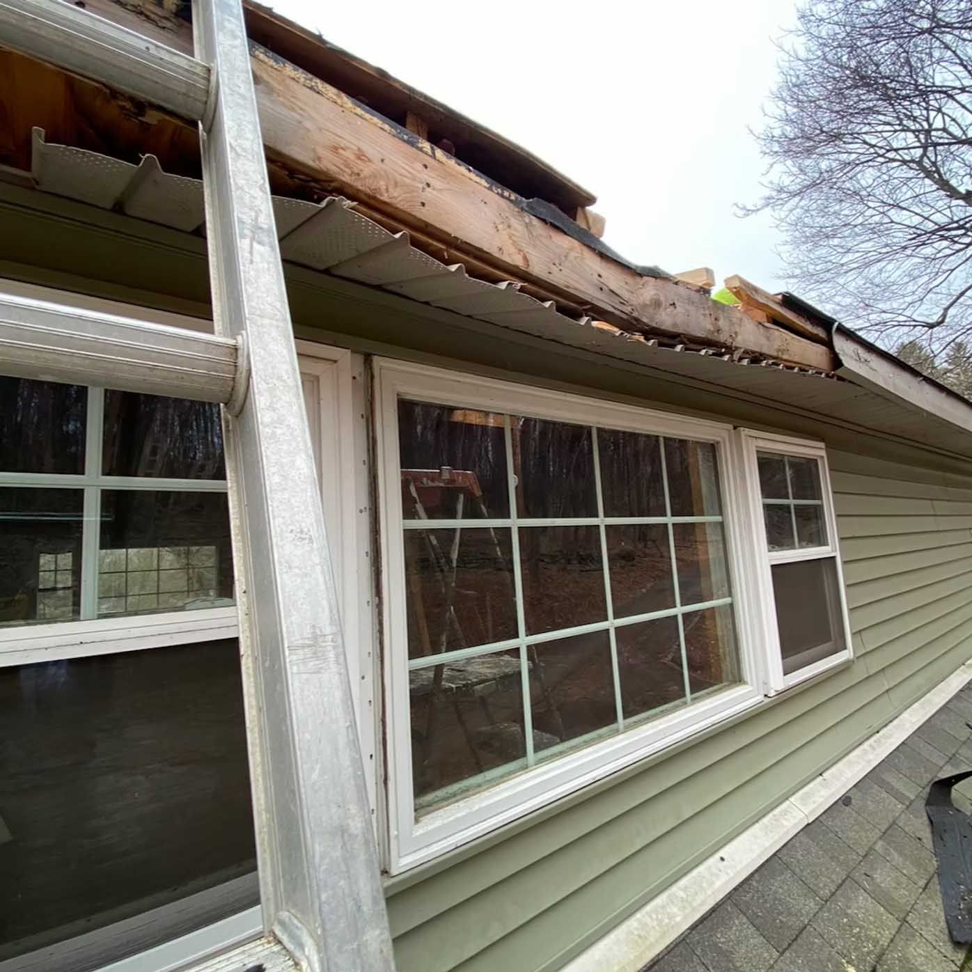 Ladder next to a house with damaged roof and windows. Green siding, white trim, and overcast sky.