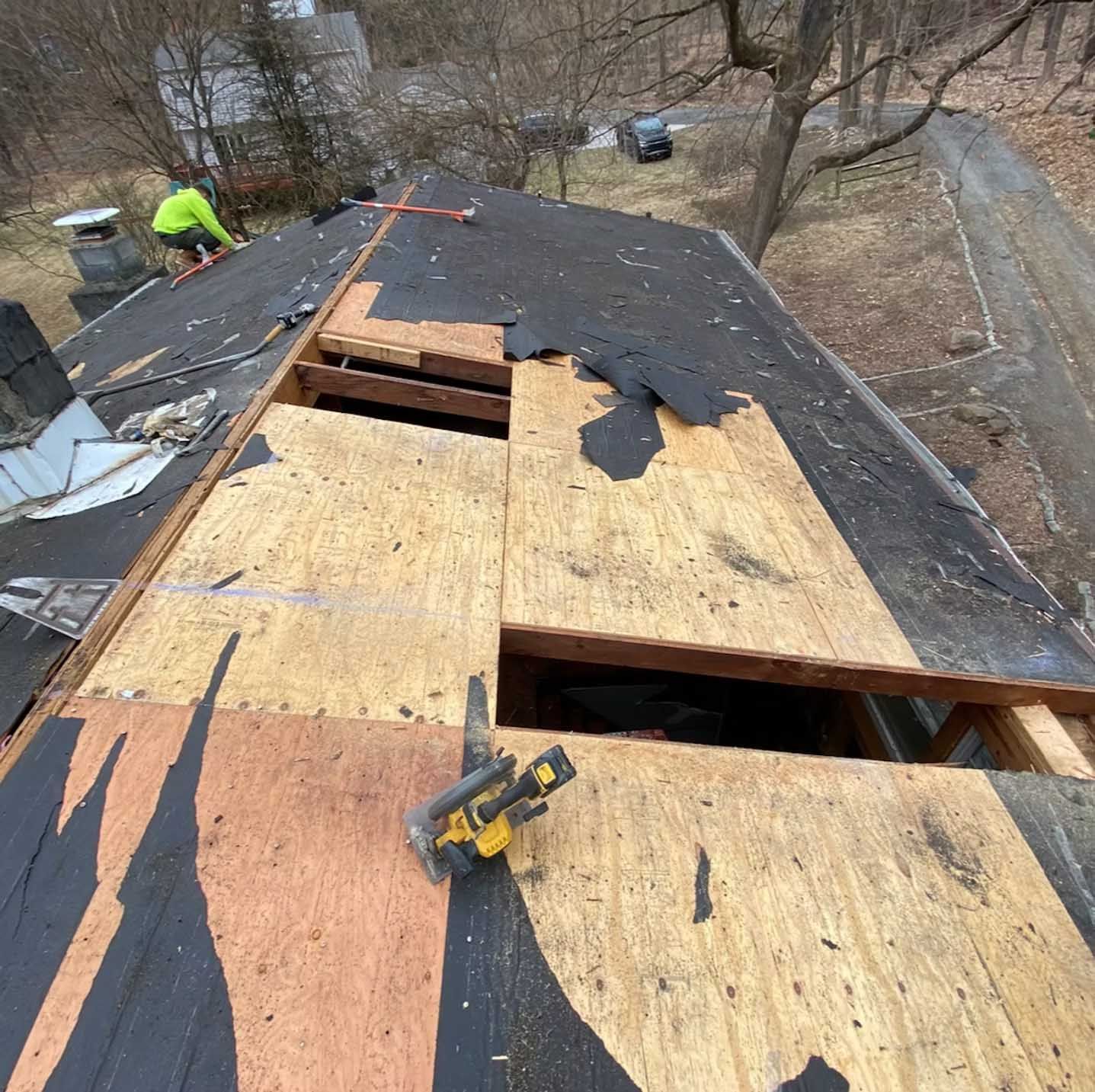 Roof being repaired, showing exposed plywood and a worker in neon vest.