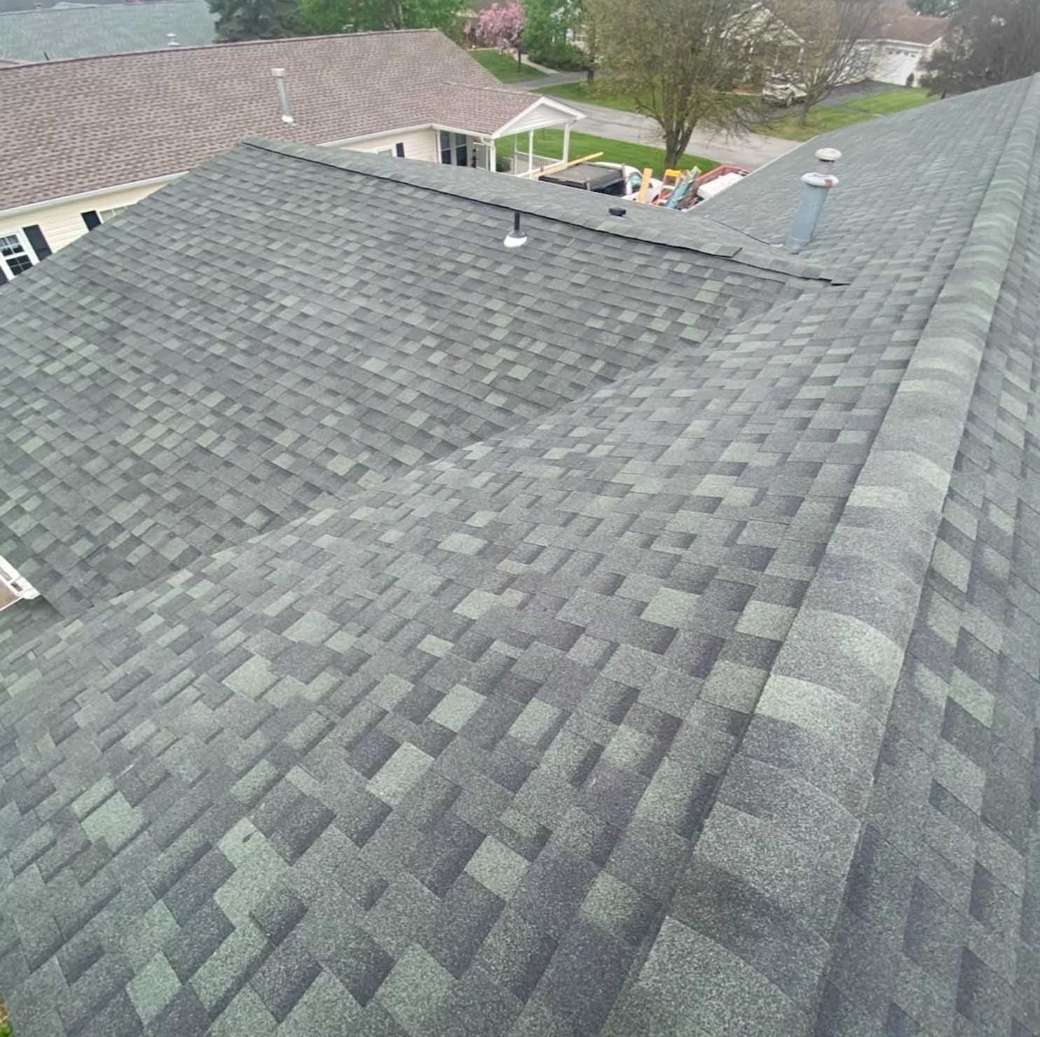 Overhead view of a gray and green asphalt shingle roof on a house, other houses visible in the background.