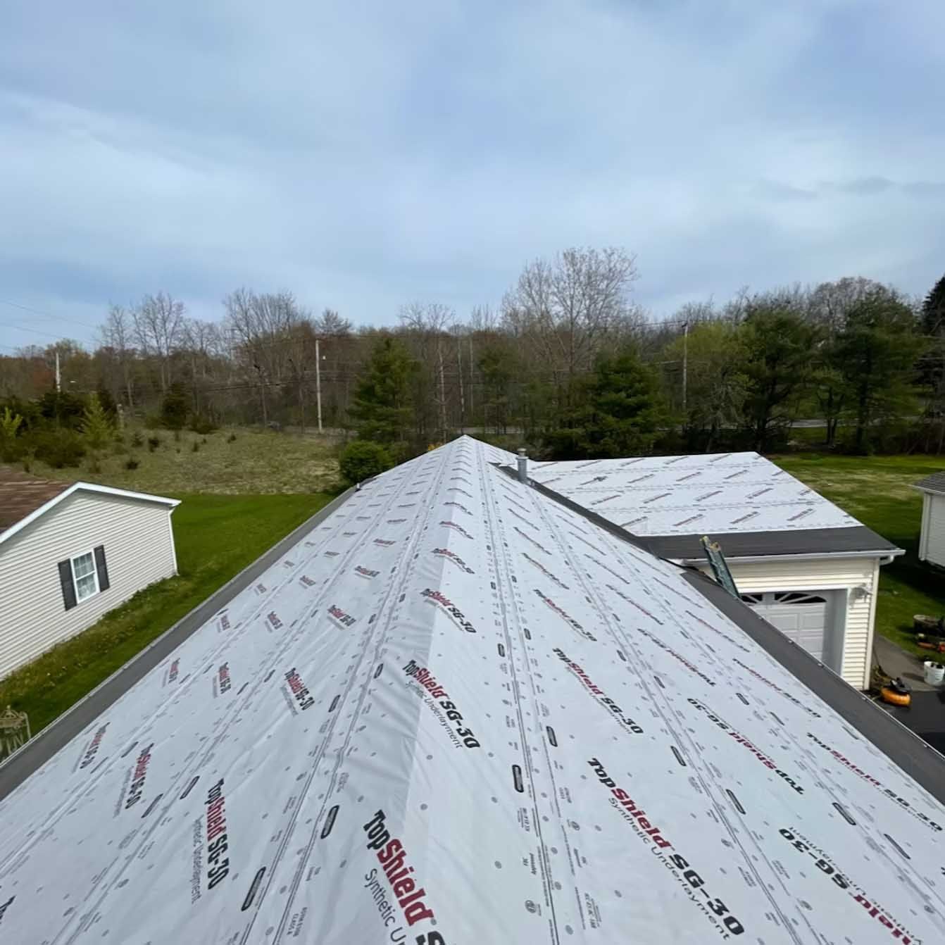 Rooftop with white underlayment in progress. House, trees, and sky in background.