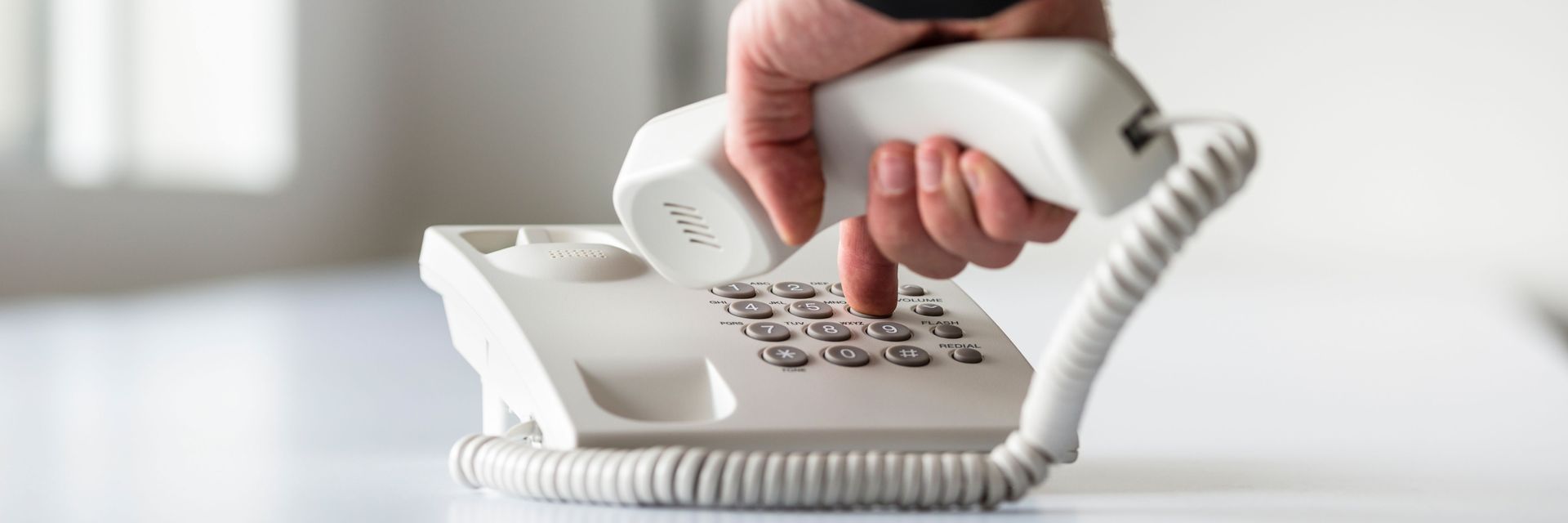 Hand holding a white corded telephone handset over the keypad on a desk
