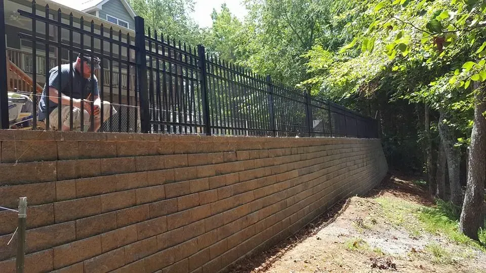 Brick retaining wall along a fenced path with a person seated on the left and trees overhead