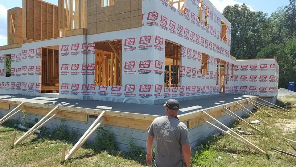 House under construction wrapped in protective house wrap, with a worker standing in front on a sunny site