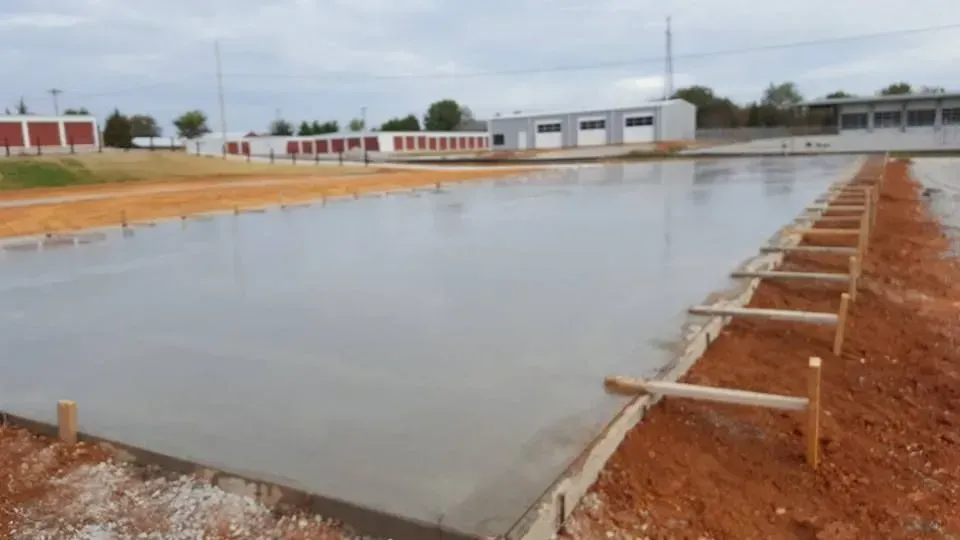 Large rectangular concrete pond under construction, with red dirt embankments and buildings in the background