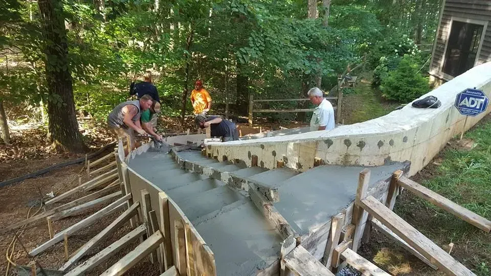 Workers pouring concrete into a large curved form in a forested outdoor construction site.