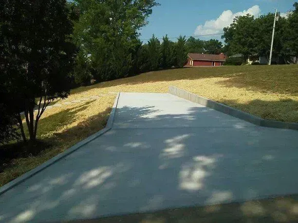 Paved walkway leading up a grassy hill beside trees and a small red building under bright sunlight