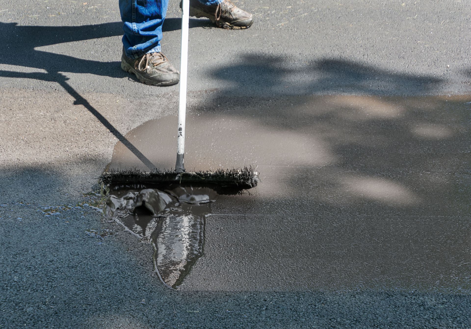 A person uses a squeegee to spread black asphalt sealer across a driveway.