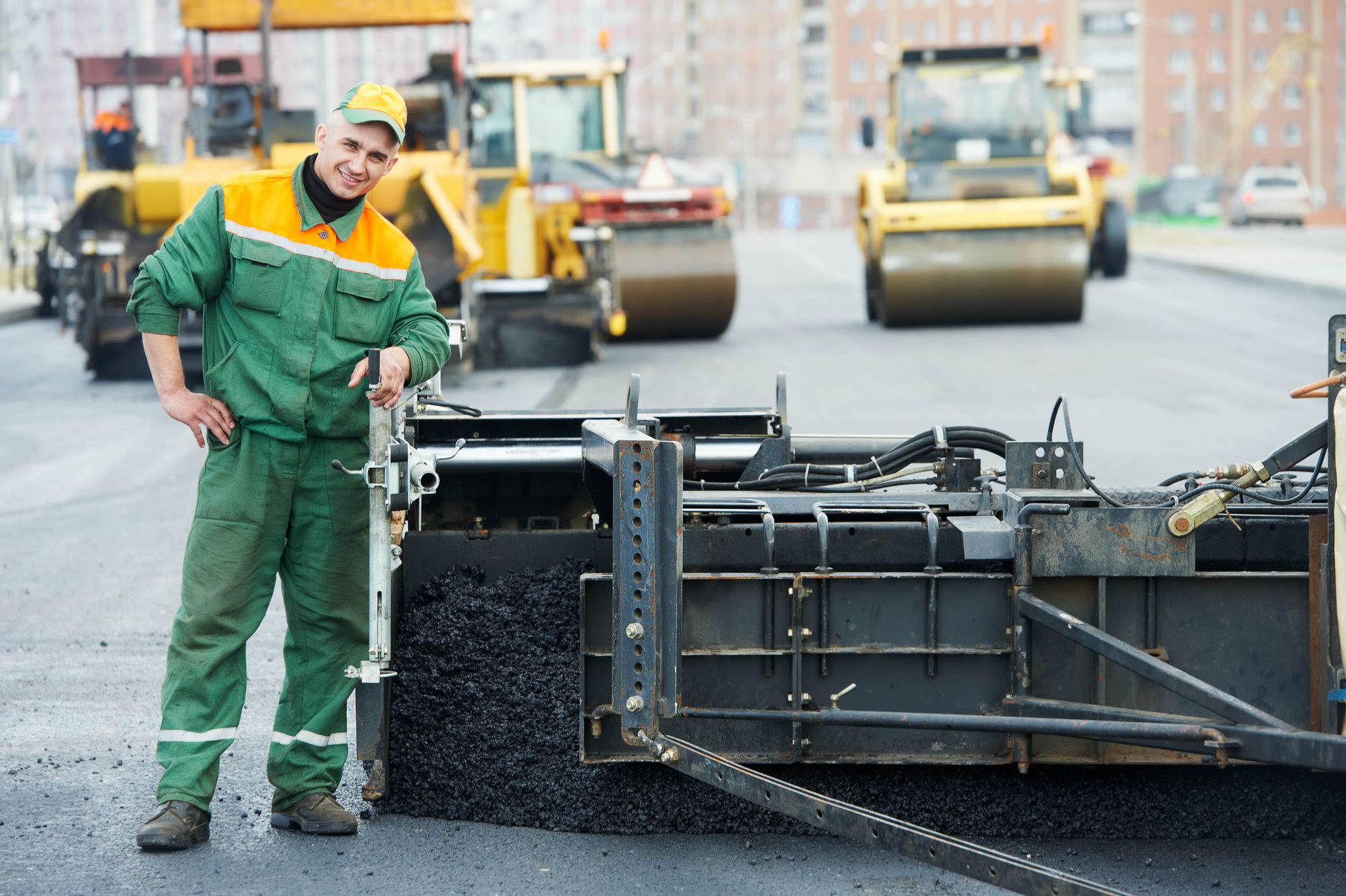 A construction worker in green and yellow gear stands next to a road paving machine on an active road construction site.
