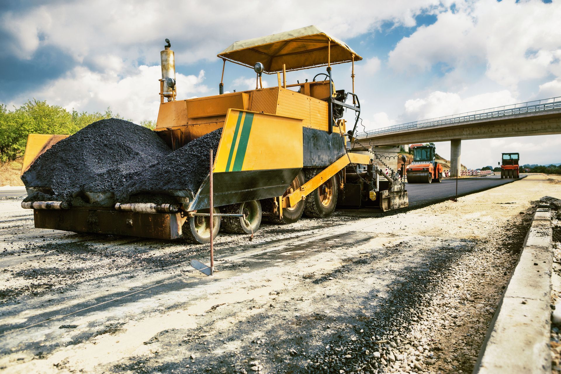 A yellow asphalt paver laying black pavement on a construction site with a bridge in the background.