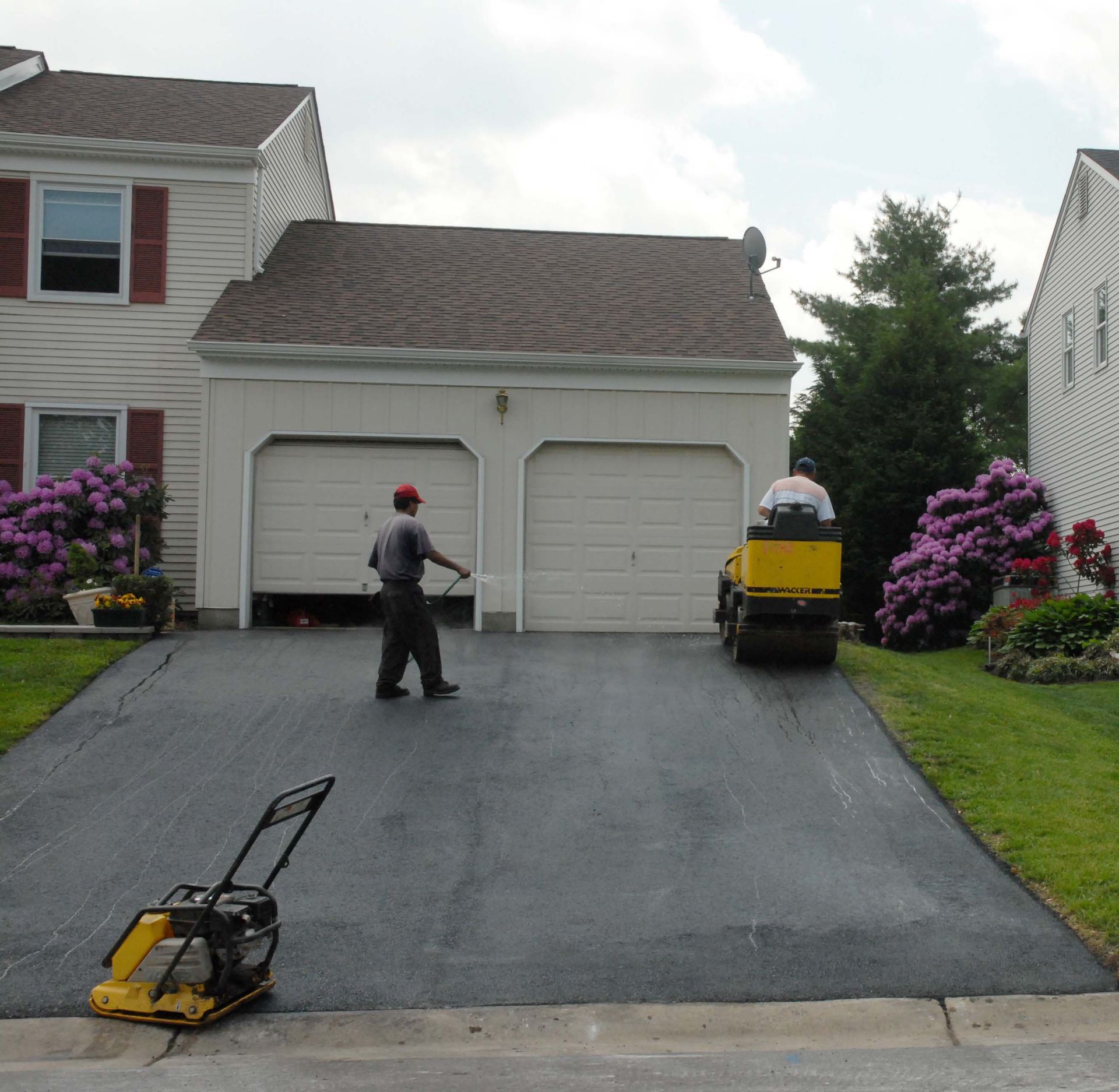 Workers pave a residential driveway with a yellow vibratory roller and a handheld plate compactor.