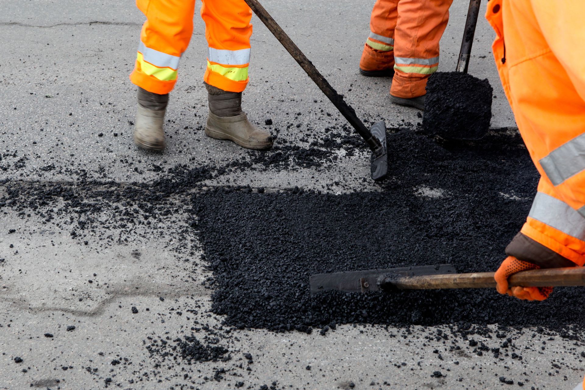 Workers in orange safety gear use shovels and tools to spread fresh asphalt on a road surface.