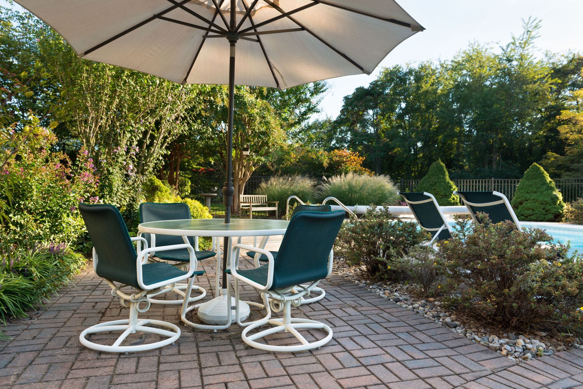 Patio set with a white umbrella, table, and chairs on a brick terrace next to a pool in a green, tree-filled backyard.