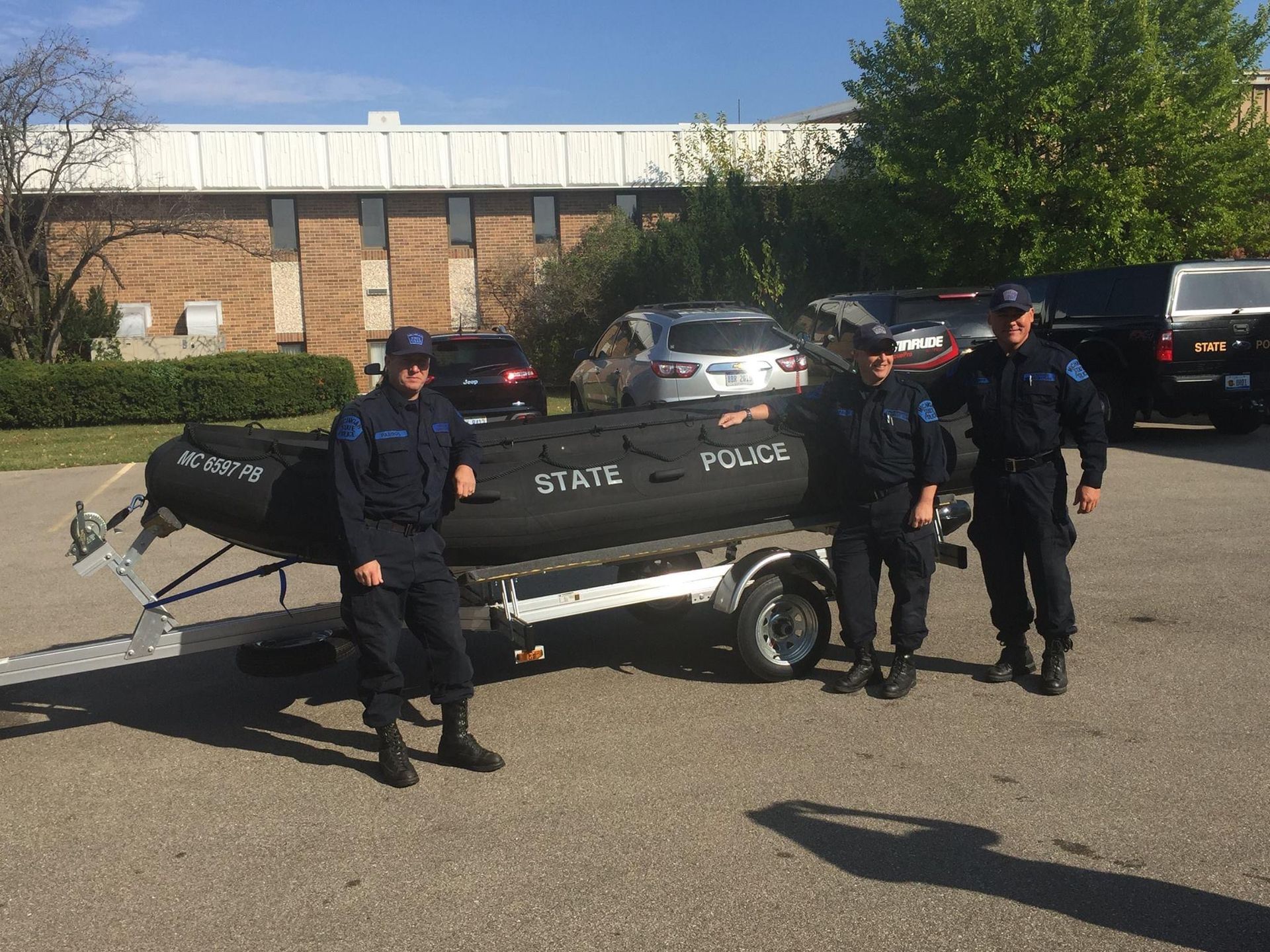 Three police officers stand beside a black inflatable boat on a trailer, labeled 