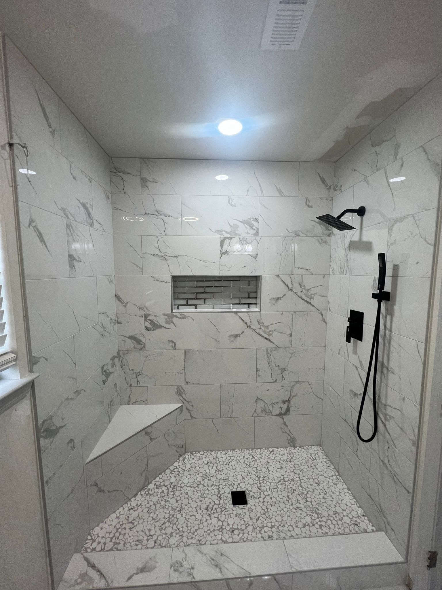Modern shower with white marble-like tiles and black fixtures, including a shower head, handheld, and built-in bench.