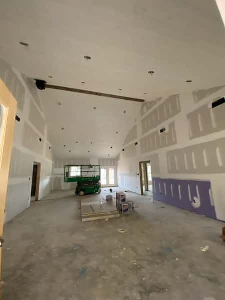 Construction site interior: unfinished walls, drywall, concrete floor, green lift, tall vaulted ceiling, brown support beam.
