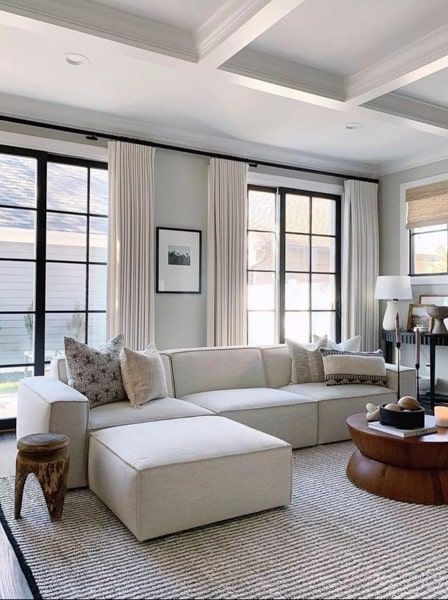 Spacious living room with cream sectional sofa, black-framed windows, white curtains, and a wooden coffee table.