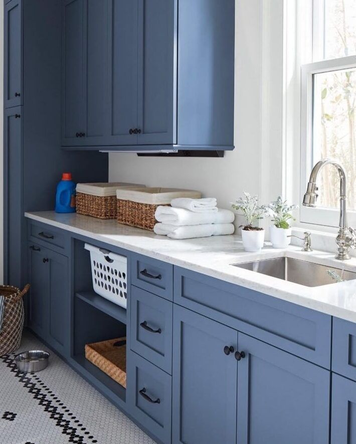 Blue laundry room with white countertops, cabinets, and stainless steel sink. Wicker baskets and laundry supplies.