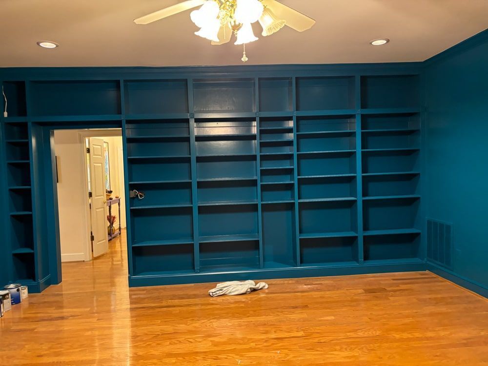 Blue built-in bookshelves and walls in a room with wooden flooring and a view into another room.