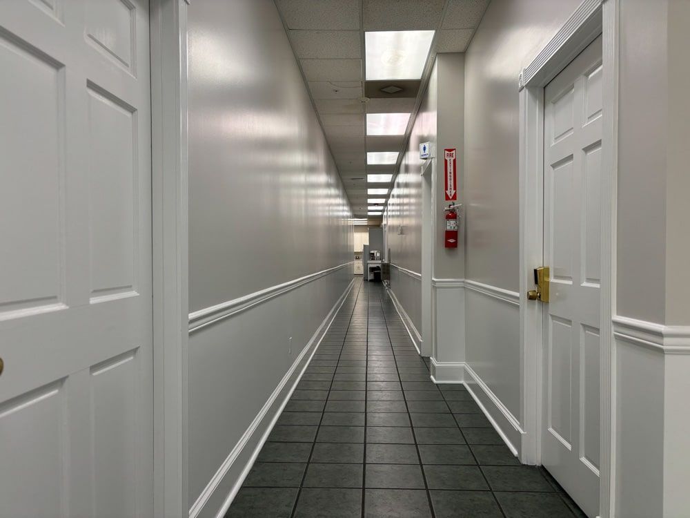 Long, narrow, light-filled hallway with doors on either side, dark tiled floor, and a fire extinguisher on the wall.