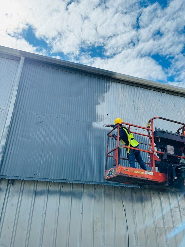Person on lift washes a corrugated metal building with a pressure washer on a sunny, cloudy day.