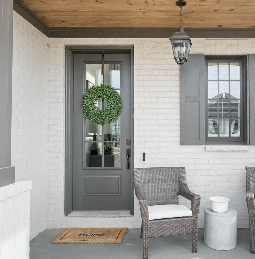 Gray front door with wreath, brick wall, shuttered window, and wicker chair on porch.