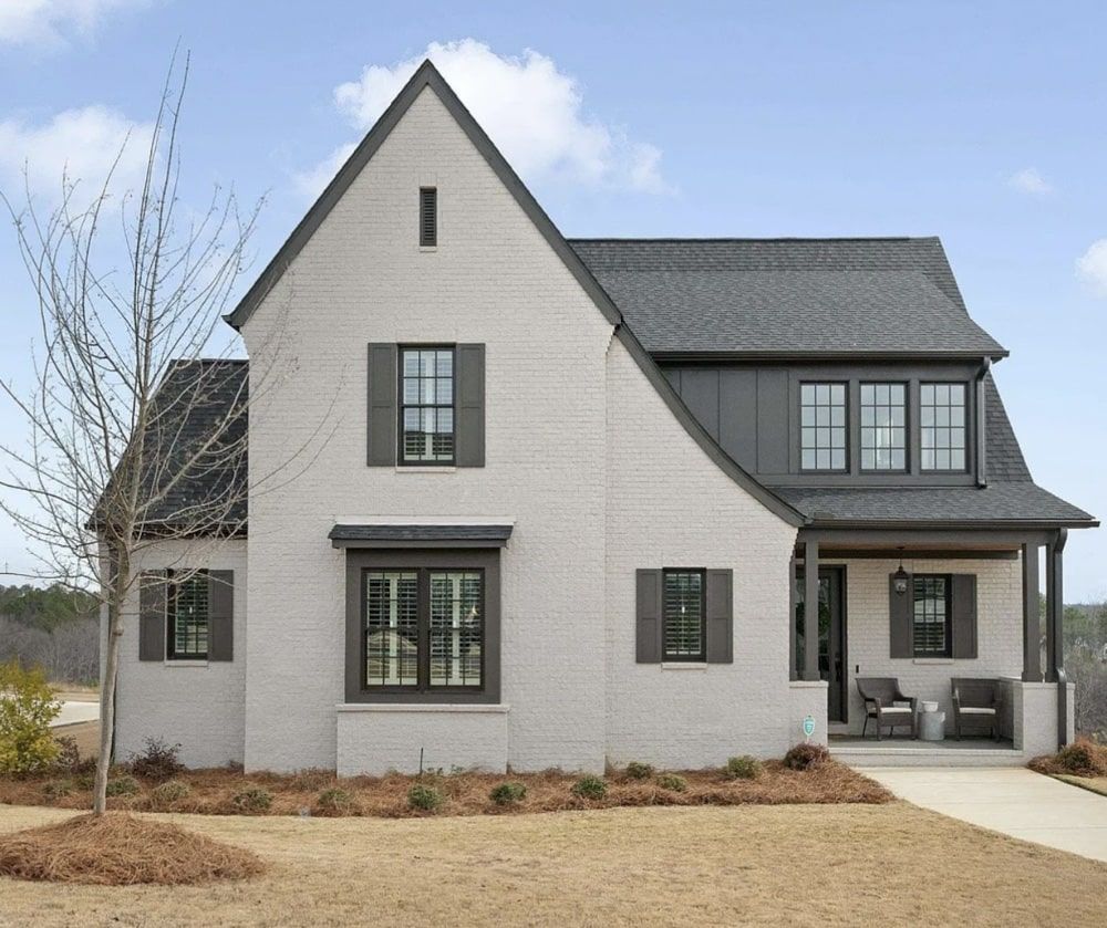 Two-story light brick house with gray roof, shutters, and trim. Porch with seating and a yard.
