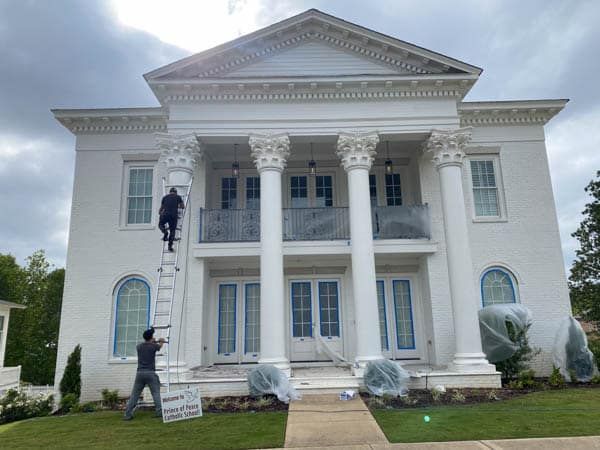 Two painters on ladders painting a white two-story house with large columns, blue doors, and covered shrubs.