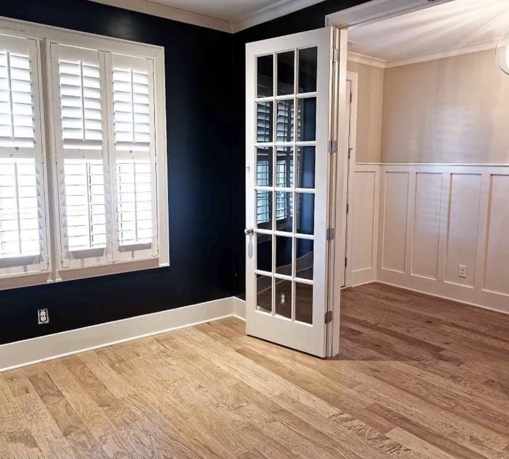 Room with hardwood floors, dark blue walls, white shutters and door, and light beige wainscoting.
