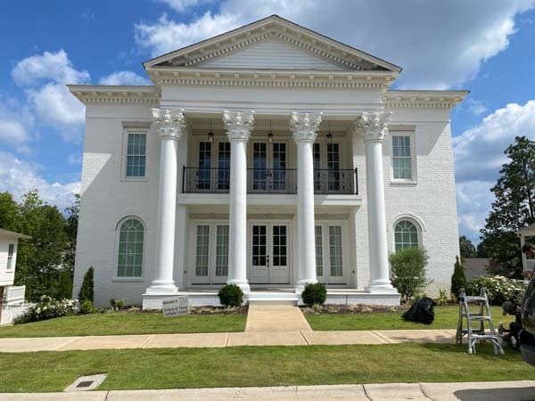 White two-story building with columns and balcony, set on a grassy lawn, blue sky.