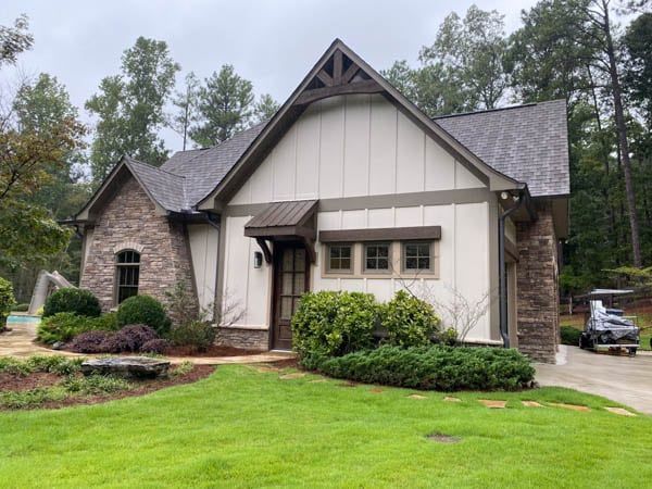 Stone and white house with brown accents, green lawn, and trees in background.