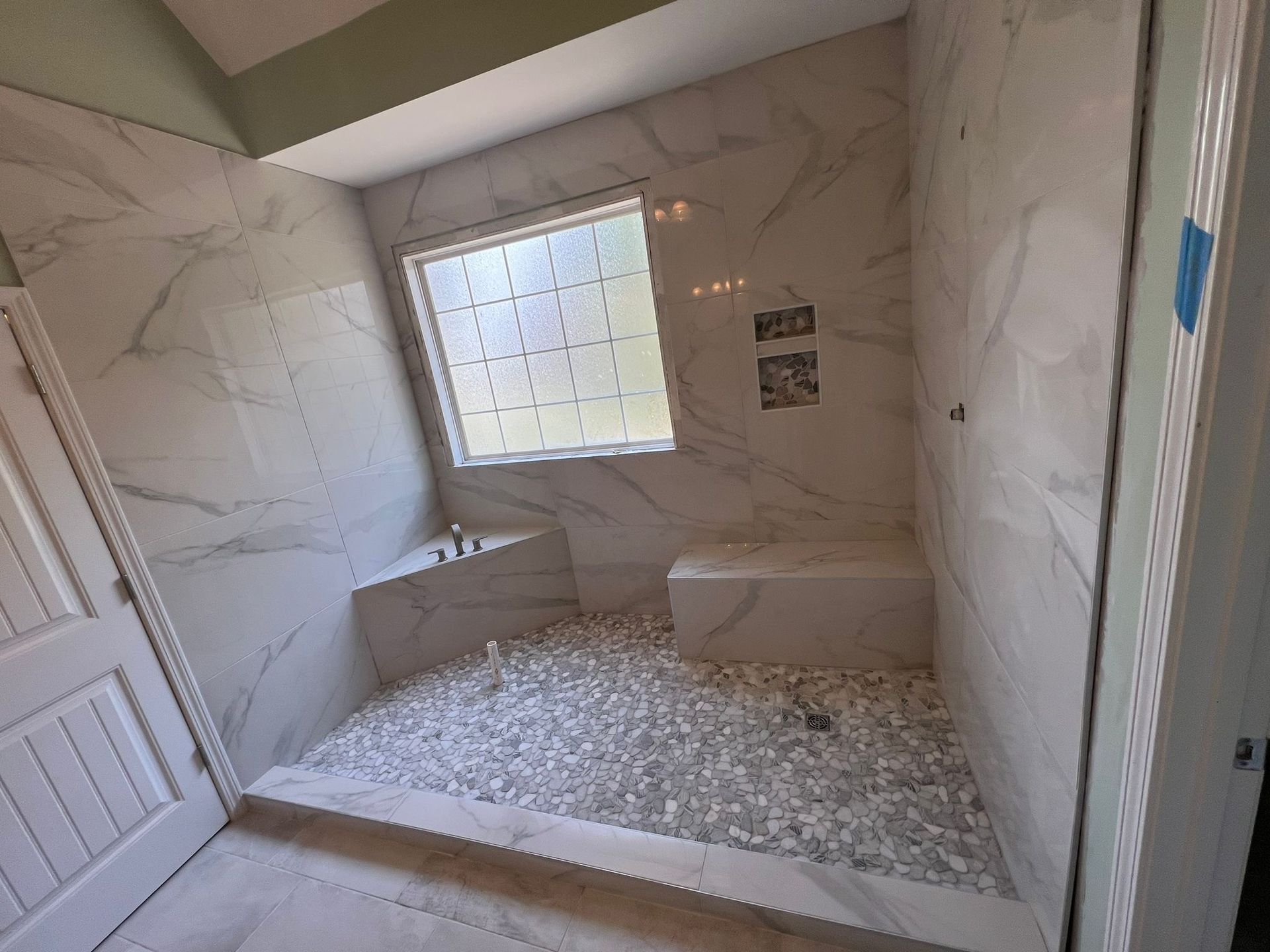 A newly renovated shower with white marble-like tiles, a window, pebble floor, and a white built-in bench.