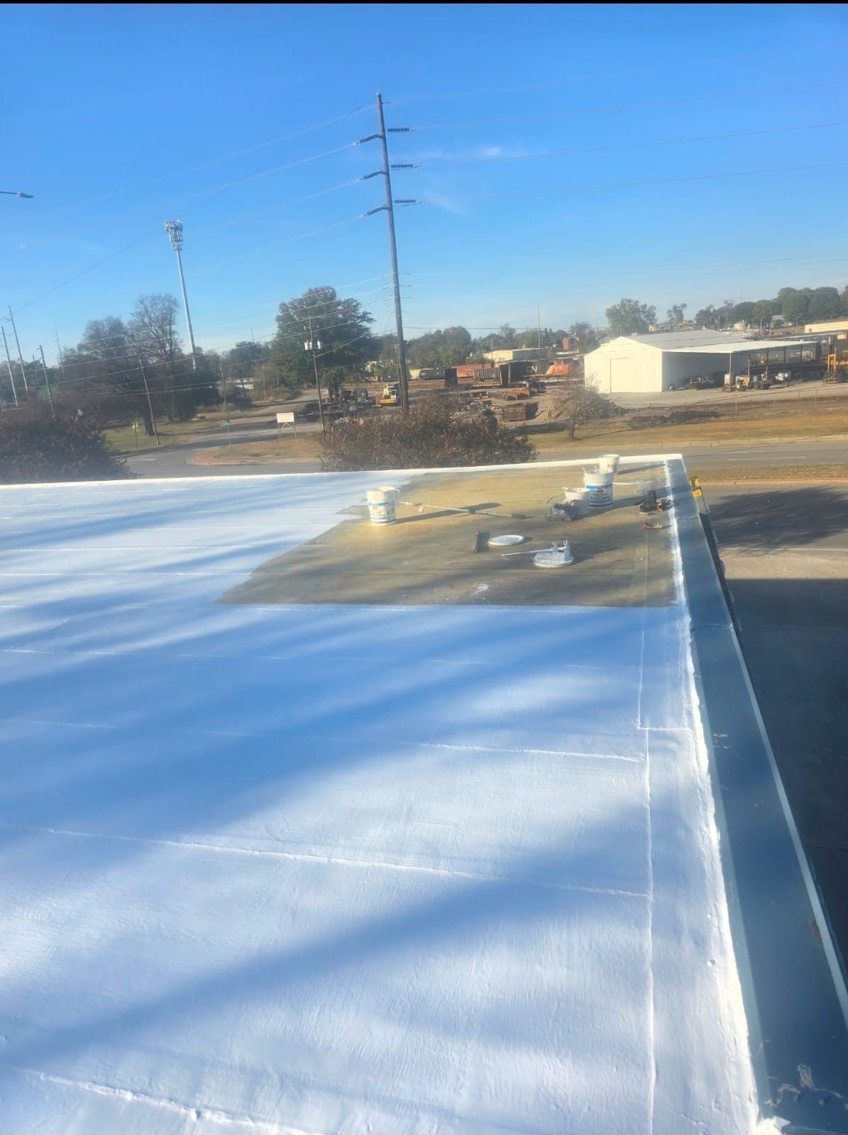 Flat commercial roof, newly coated white, in sunlight. Buckets and equipment visible. Distant trees and buildings.