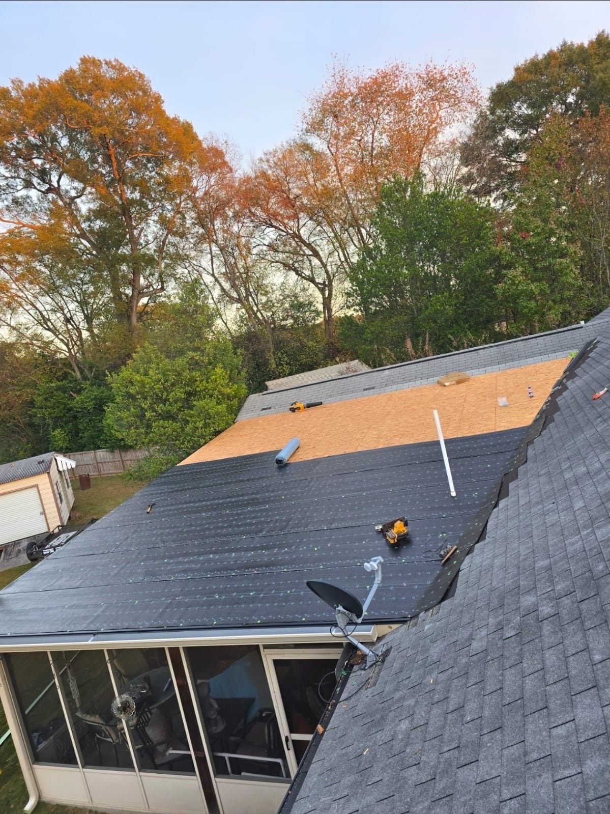 Roof partially covered with new shingles and plywood; tools on the roof; trees in background.